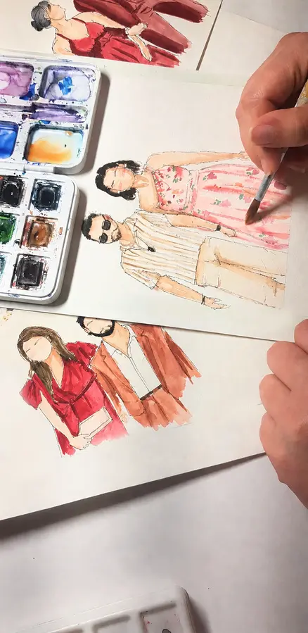 Tessa peint un couple à l’aquarelle, sa palette ouverte à côté des portraits déjà terminés